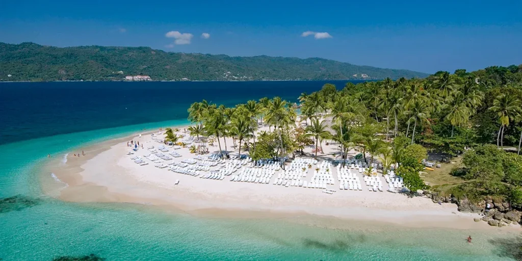 Vista aérea panorámica de la isla Cayo Levantado en la Bahía de Samaná, República Dominicana. Una playa de arena blanca curvada está poblada por cientos de tumbonas de playa blancas y ordenadas, y flanqueada por una densa arboleda de palmeras. Las aguas turquesas cristalinas y azules profundas rodean la isla, con montañas y un resort visible al fondo bajo un cielo azul claro