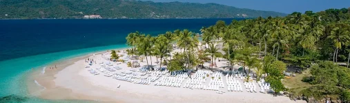 Vista aérea panorámica de la isla Cayo Levantado en la Bahía de Samaná, República Dominicana. Una playa de arena blanca curvada está poblada por cientos de tumbonas de playa blancas y ordenadas, y flanqueada por una densa arboleda de palmeras. Las aguas turquesas cristalinas y azules profundas rodean la isla, con montañas y un resort visible al fondo bajo un cielo azul claro